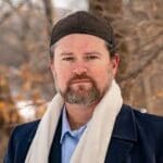 Bearded man wearing a dark knit cap and cream scarf, outdoors in a park with bare trees in autumn.
