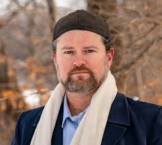 Bearded man wearing a dark knit cap and cream scarf, outdoors in a park with bare trees in autumn.