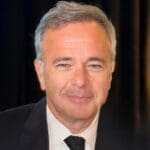 Smiling man in a black suit and tie, head-and-shoulders portrait against a dark background.