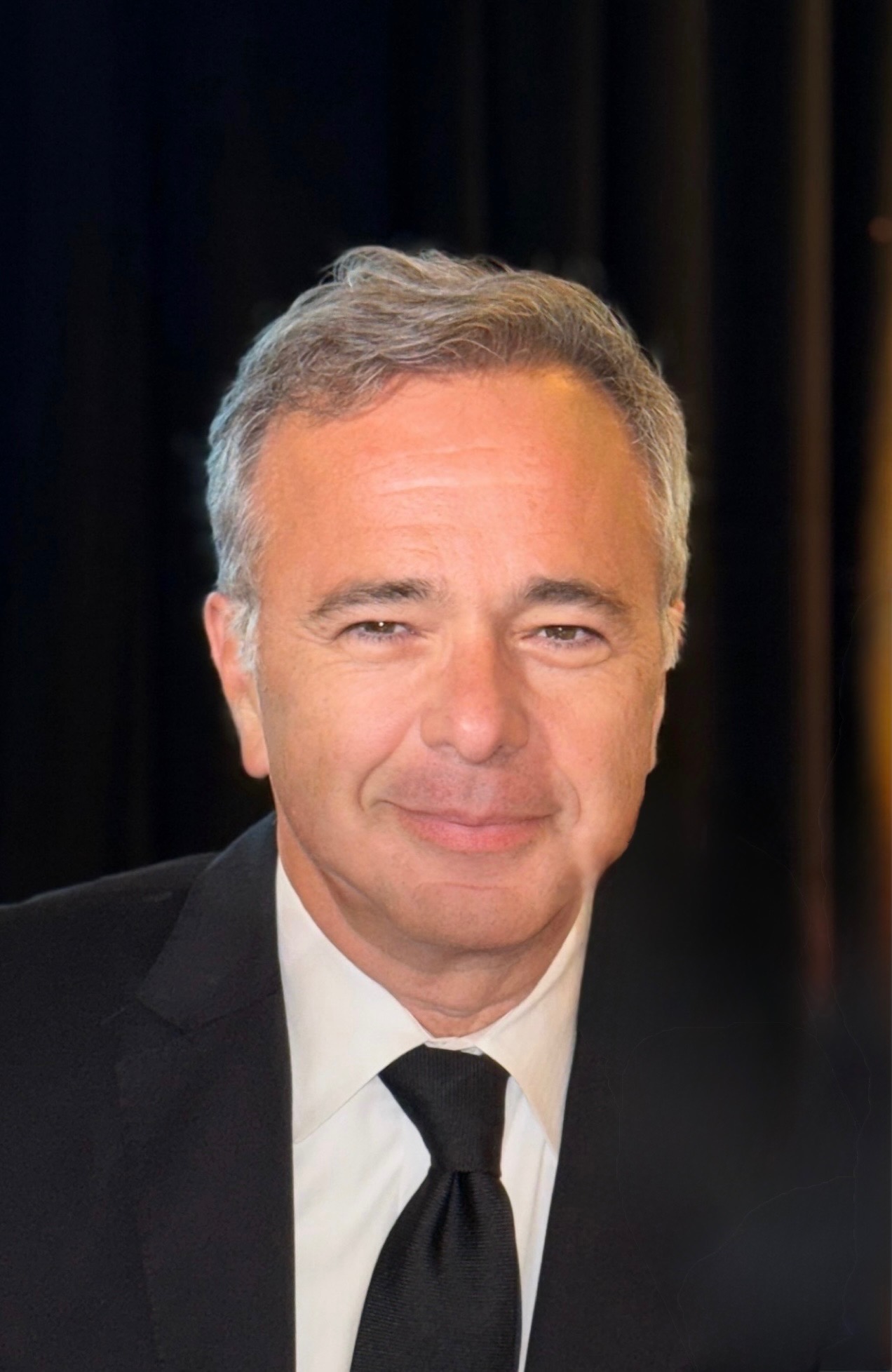 Smiling man in a black suit and tie, head-and-shoulders portrait against a dark background.