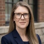 Professional headshot of a woman with tortoiseshell glasses and a navy blazer, standing outdoors.