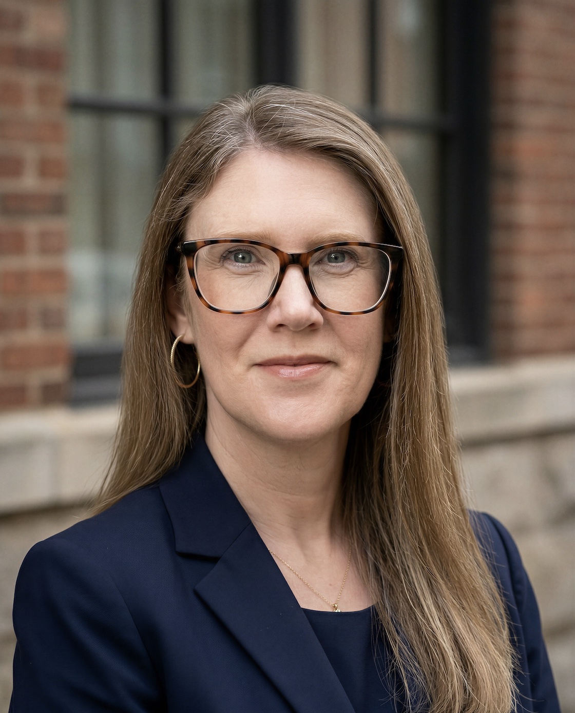 Professional headshot of a woman with tortoiseshell glasses and a navy blazer, standing outdoors.